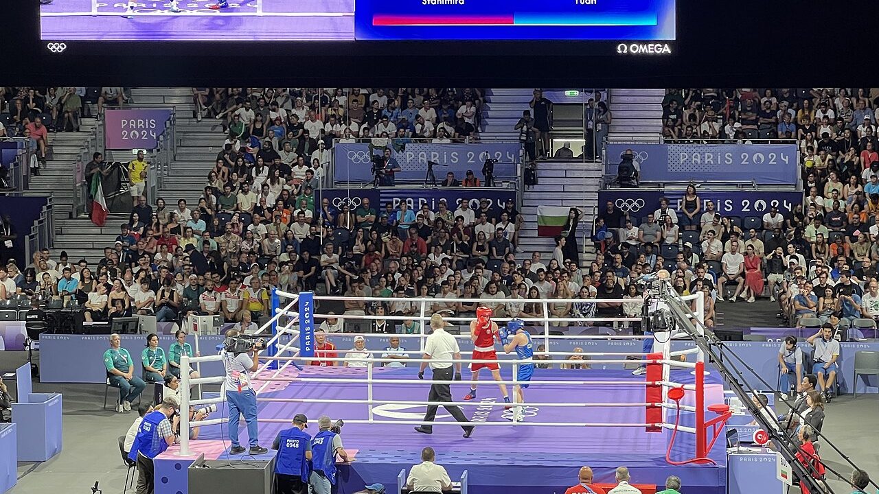 ภาพมุมกว้างของสนามแข่งมวยสากลโอลิมปิกปารีส 2024 เห็นเวทีกลางสนาม โต๊ะกรรมการรอบเวที และจอแสดงผลด้านบน