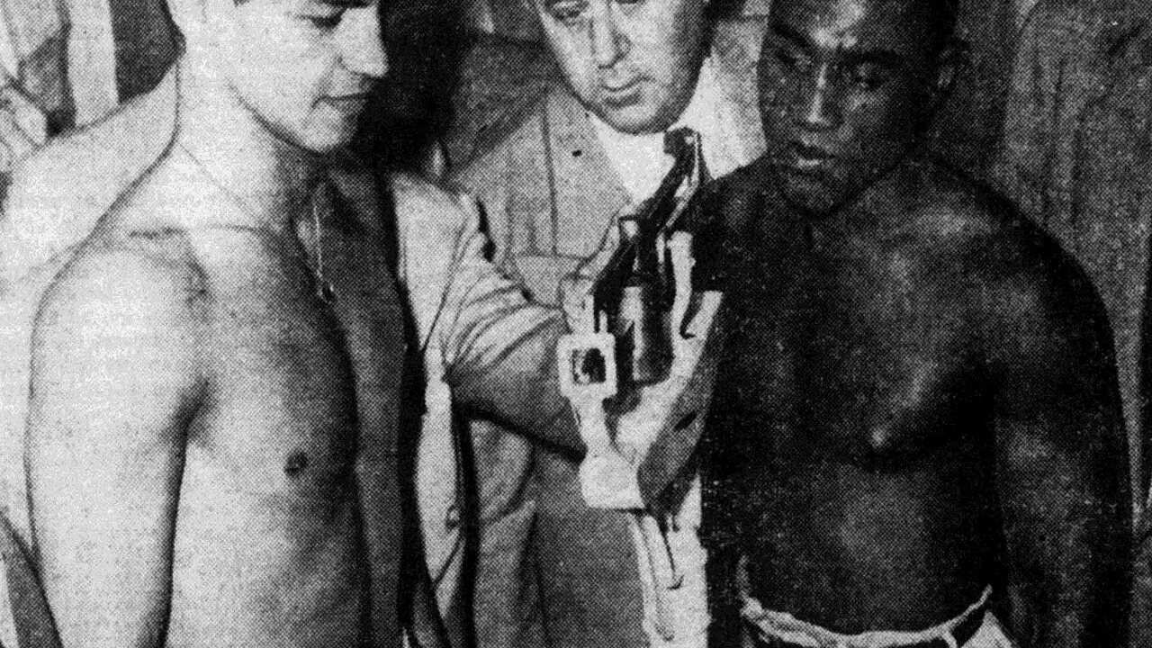 ภาพถ่ายขาวดำปี 1951 นักมวยสองคนยืนเปลือยท่อนบนข้างเครื่องชั่งน้ำหนัก โดยมีเจ้าหน้าที่ยืนอ่านตัวเลขอยู่ตรงกลาง