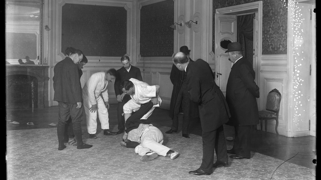 การสาธิตยูยิตสูในห้องโถงที่กองบัญชาการตำรวจกรุงปารีส ปี 1905 ผู้สาธิตกดคู่ฝึกลงกับพื้นโดยมีชายสวมสูทและหมวกยืนล้อมดู