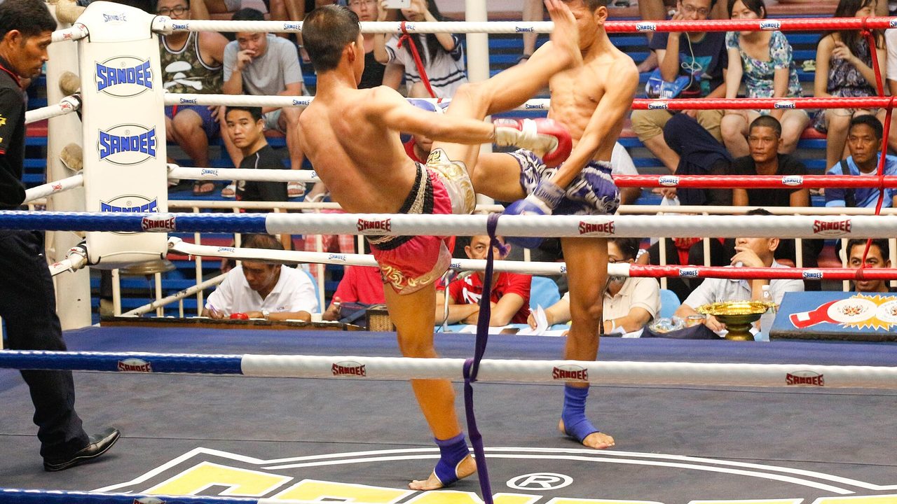 การแข่งขันมวยไทยบนเวทีในร่ม นักมวยสองคนปะทะกันกลางเวทีโดยมีผู้ชมล้อมรอบ