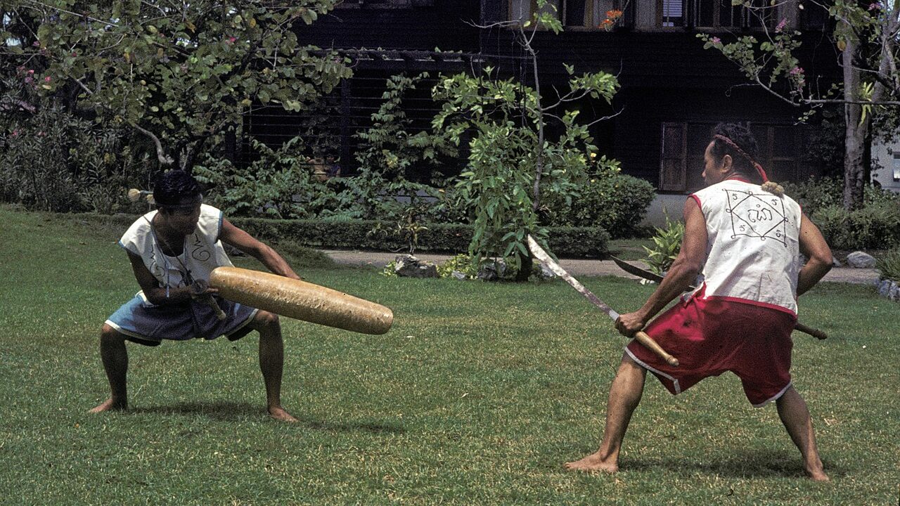 นักแสดงกระบี่กระบองสองคนในกรุงเทพฯ ปี 1965 คนหนึ่งถือดั้งทรงยาวและอาวุธสั้น อีกคนถือดาบ ทั้งคู่ย่อเข่าเข้าหากันบนสนามหญ้า