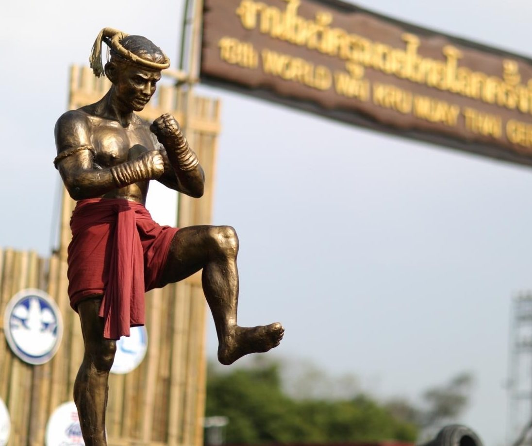รูปปั้นจำลองนายขนมต้มสีทองนุ่งผ้าสีแดง ยืนยกเข่าตั้งการ์ด ด้านหลังมีป้ายงานไหว้ครูมวยไทย