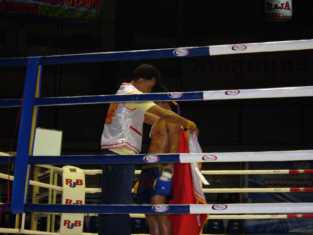 ภาพการชกมวยไทยบนเวทีในสนามมวยราชดำเนิน มีกรรมการยืนคุมอยู่กลางเวทีและผู้ชมเต็มอัฒจันทร์โดยรอบ