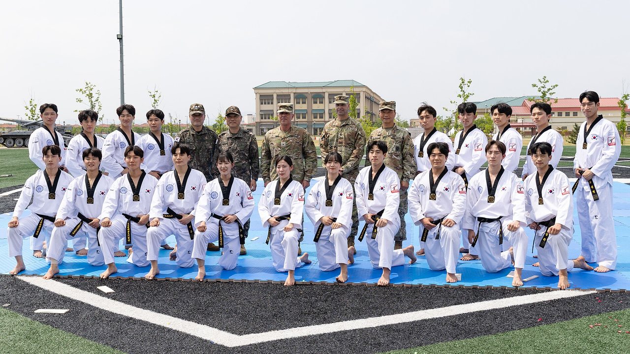 ทีมเทควันโดสาธิตท่าพร้อมกันเป็นแถวในโรงยิม โดยมีผู้ชมนั่งดูอยู่รอบ