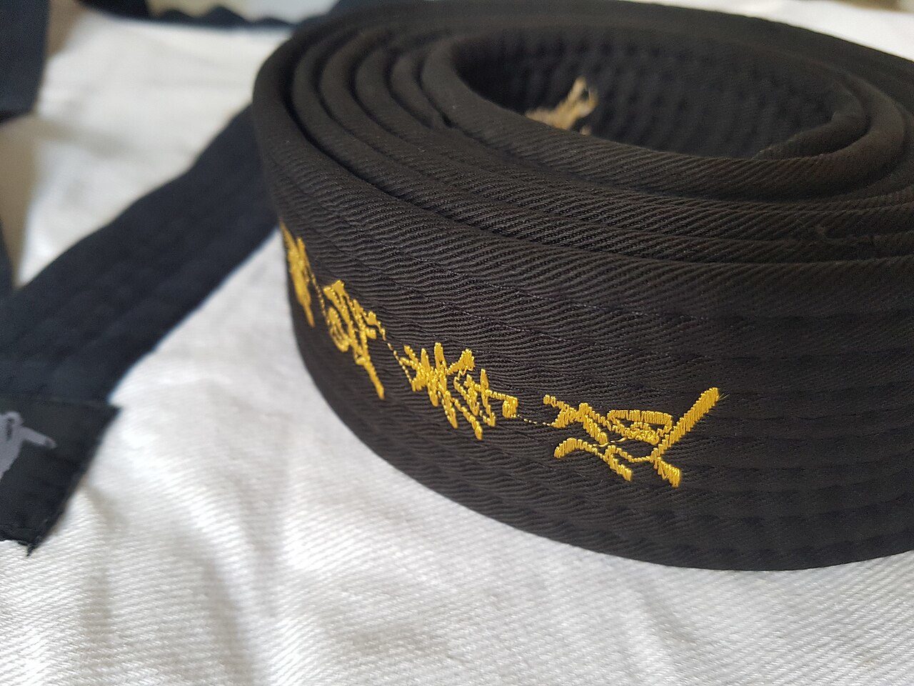 สายดำเทควันโดม้วนวางอยู่บนชุดโดบกสีขาว ปลายสายปักตัวอักษรด้วยด้ายสีทอง